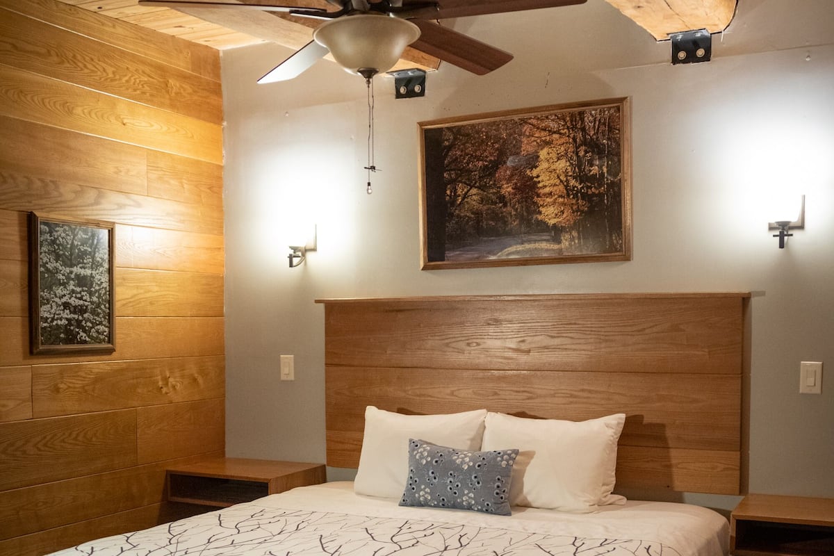 A bedroom with a wooden ceiling, a bed with a white bedspread, a ceiling fan, and a painting on the wall.