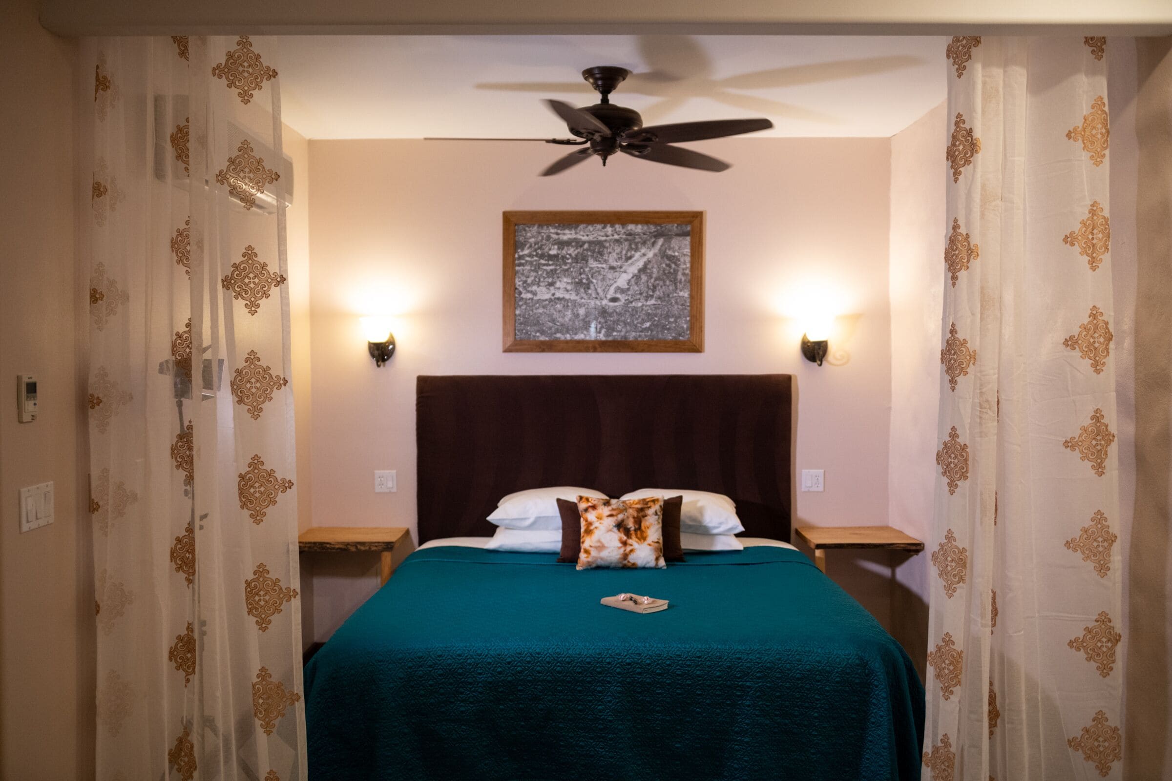 Una cama bien hecha con una colcha color turquesa, dos almohadas y una almohada decorativa. Arriba, un ventilador de techo, dos lámparas de noche y un cuadro en la pared.