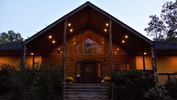 A rustic lodge with a steeply pitched roof, a central entrance with a door, and a porch with multiple lights. - Makanda Inn (Makanda)