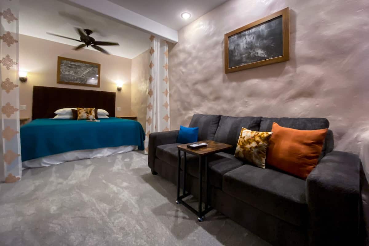 A hotel room with a bed, a sofa, a small table, a ceiling fan, and a painting on the wall.