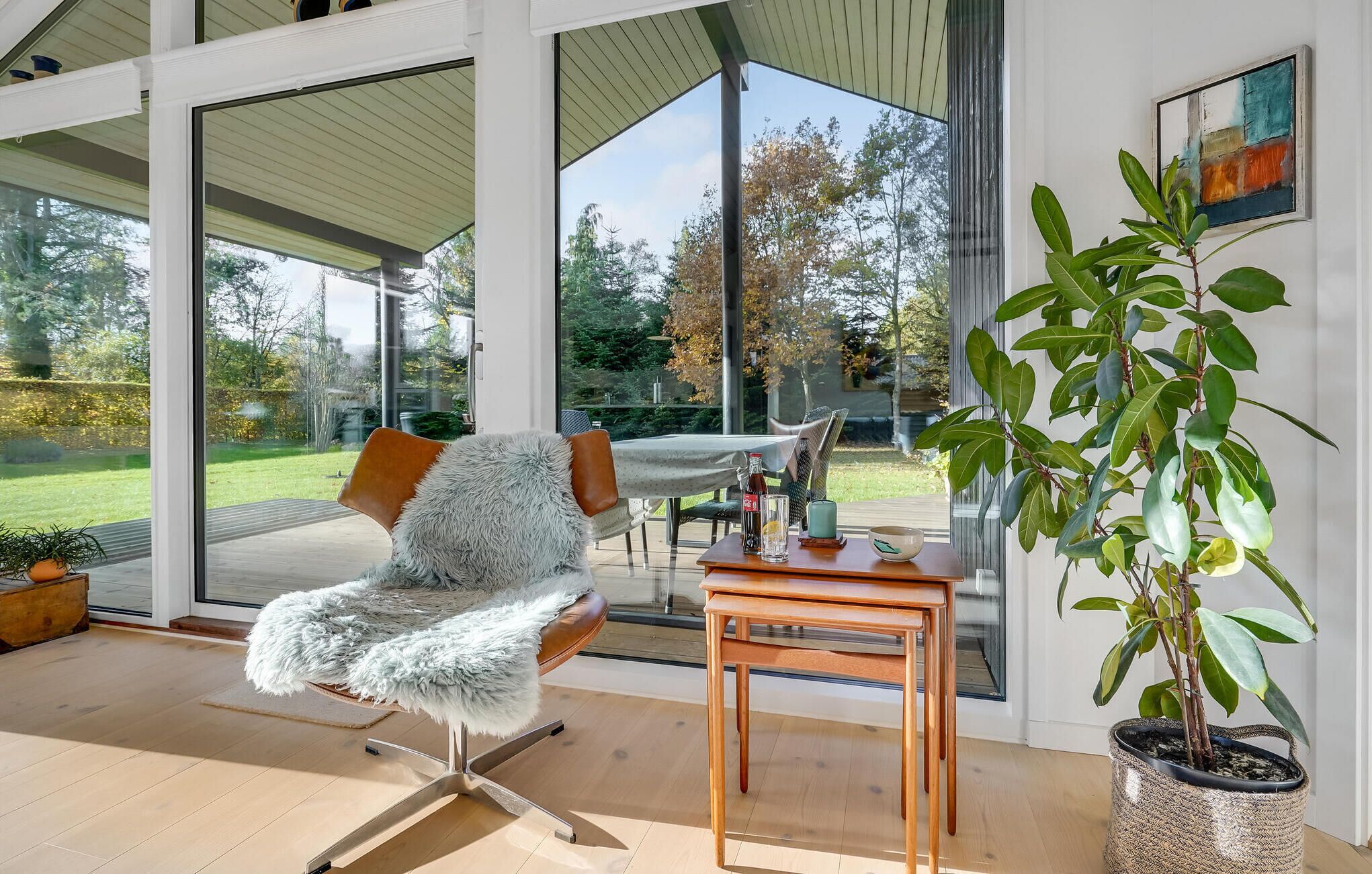 Un interior moderno con un ventanal que da a un jardín, un sillón mullido, una mesa de madera y una planta en maceta.