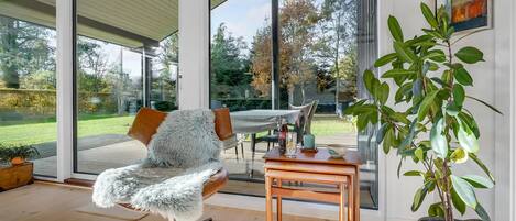 Un interior moderno con un ventanal que da a un jardín, un sillón mullido, una mesa de madera y una planta en maceta.
