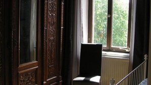 Une pièce comprenant une grande armoire en bois, une chaise, une fenêtre donnant sur un paysage verdoyant et une rambarde blanche.