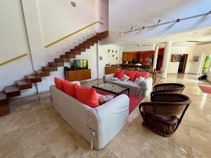 A spacious living area with a staircase, a sofa with red cushions, a coffee table, wicker chairs, and a kitchen in the background. - Contemporary Villa in Lush, Tropical, Private Paradise (Bucerias)