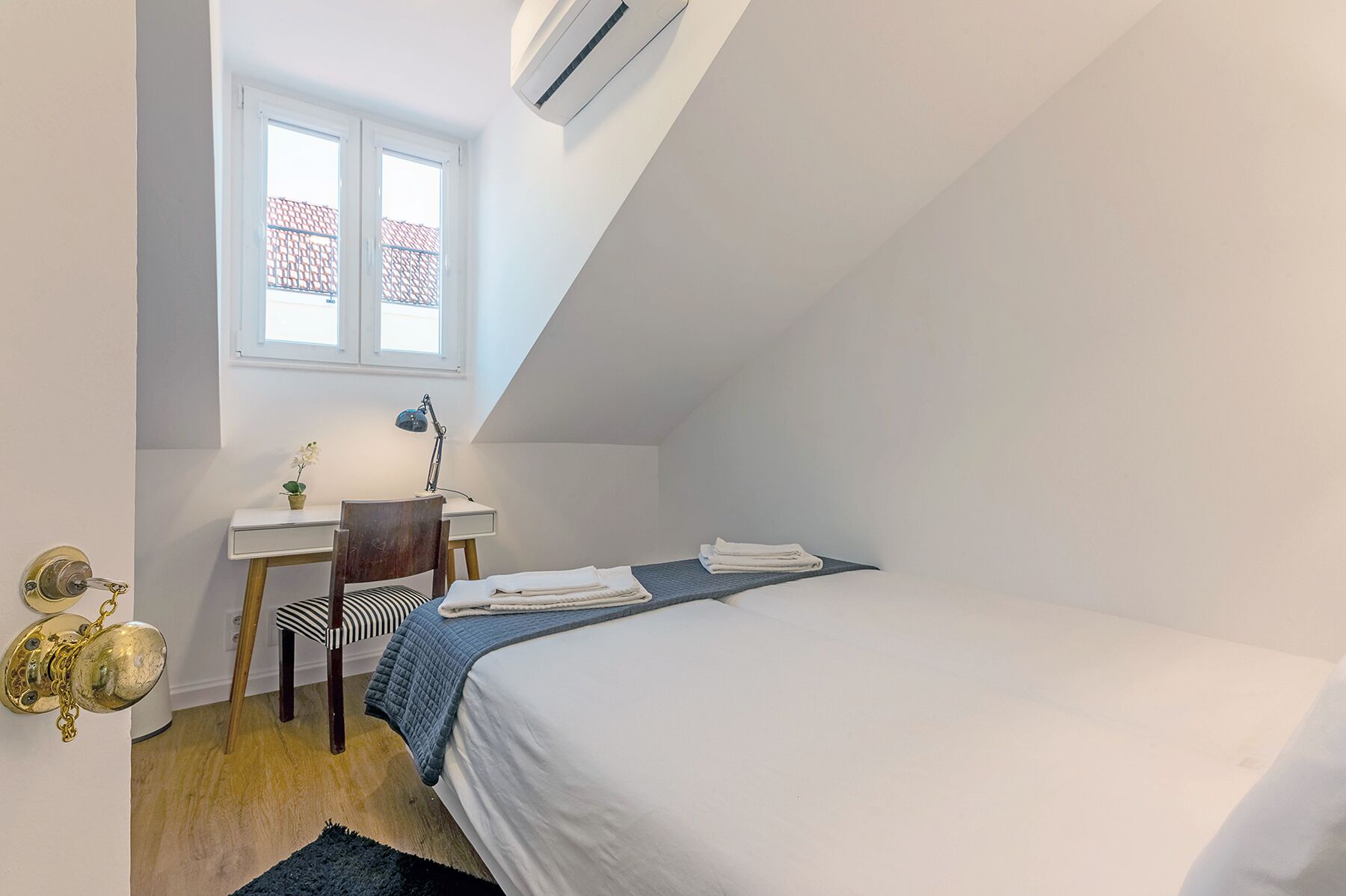 A small, tidy room with a bed, a desk, a chair, a window, and an air conditioning unit.