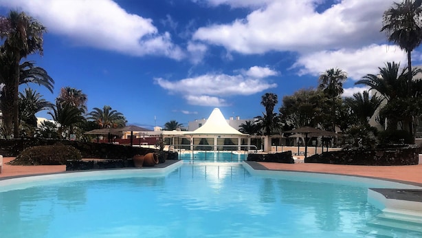 A large swimming pool with a white gazebo in the background, surrounded by palm trees and a clear sky. - Top floor apartment with amazing ocean views  (Costa Teguise)