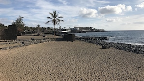 A beach with a palm tree, rocky shoreline, and buildings in the background. - Top floor apartment with amazing ocean views  (Costa Teguise)