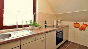 Una cucina con finestra, lavello, piano di lavoro, piante in vaso, bottiglia di vino, forno e presine appese al muro.