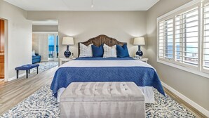 A bedroom with a large bed, two bedside lamps, a ceiling fan, and a view of the ocean through the window.