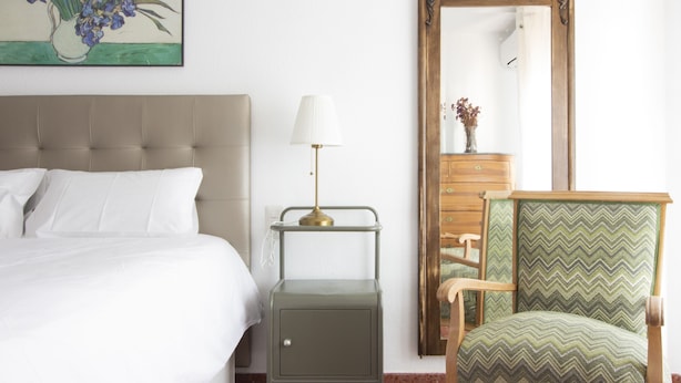 Ein Hotelzimmer mit einem Bett, einem Nachttisch mit Lampe, einem Spiegel, einer Kommode und einem Sessel.