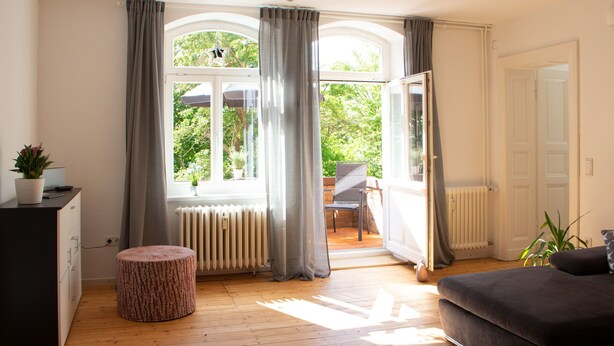 A room with a wooden floor, a grey sofa, a small round ottoman, a radiator, and a view through a window with curtains. - Hufelandstern - Beautiful old building vacation apartment for up to 7 people with e-charging station (Bad Wildungen)