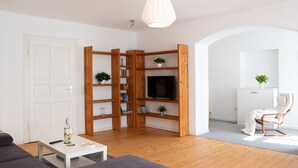 A modern living room with a wooden bookshelf, a flat-screen TV, a white door, and a seating area with a sofa and a chair. - Hufelandstern - Beautiful old building vacation apartment for up to 7 people with e-charging station (Bad Wildungen)