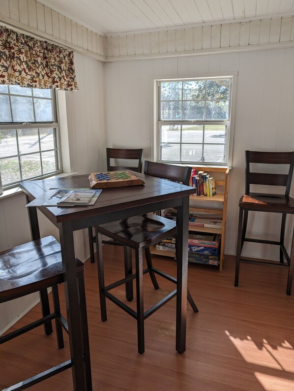 A dining area with a wooden table and chairs, a window with a view of trees, and a bookshelf with board games.