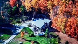 A modern house with a large deck, surrounded by a forest with autumn foliage. - Cherry-Pick the Ideal Getaway!  4BR/3BA home @ Traverse City & Torch Lake (Williamsburg)