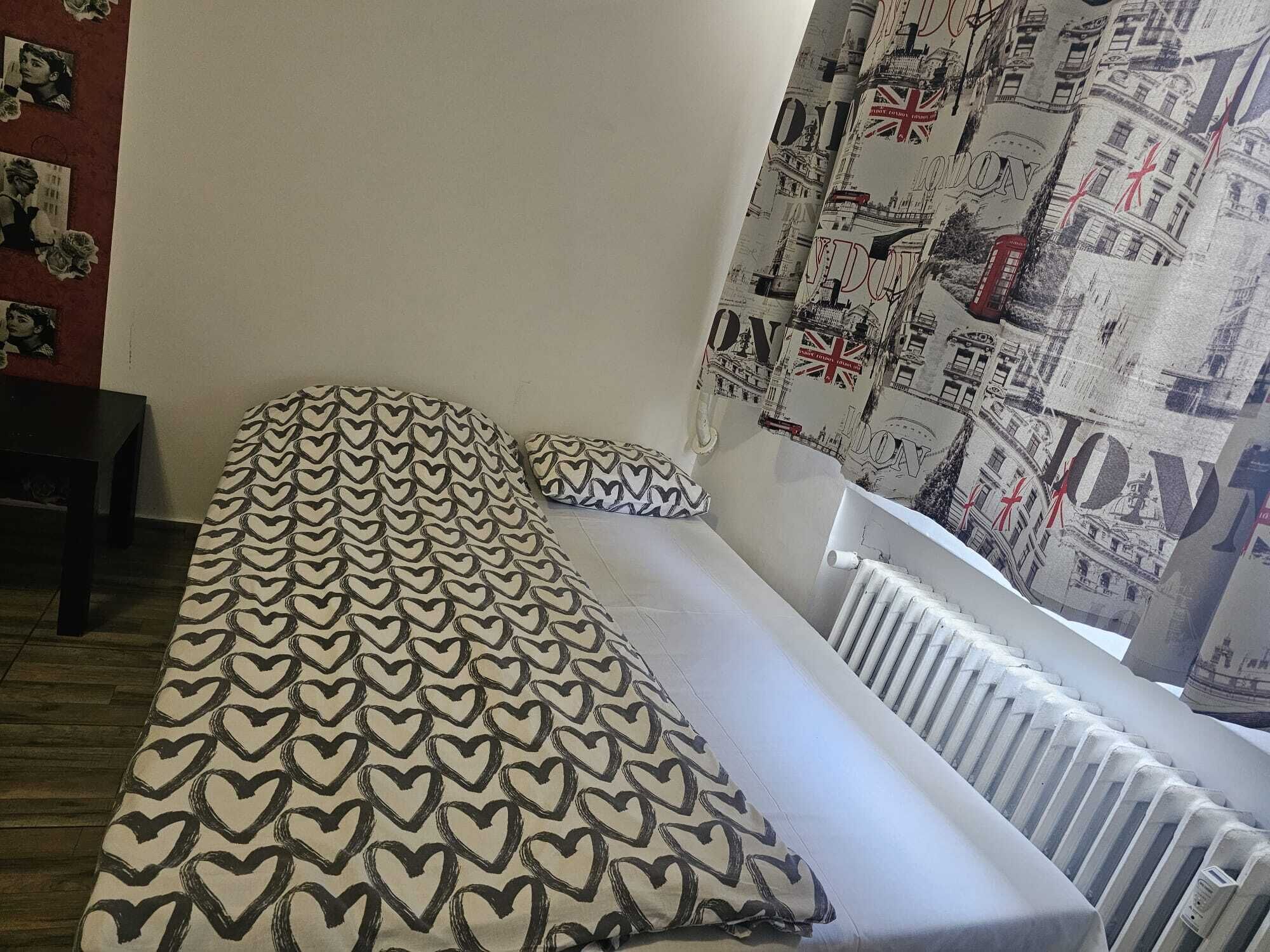 A single bed with a heart-patterned bedspread and a pillow, a radiator, and a wall with a London-themed poster.