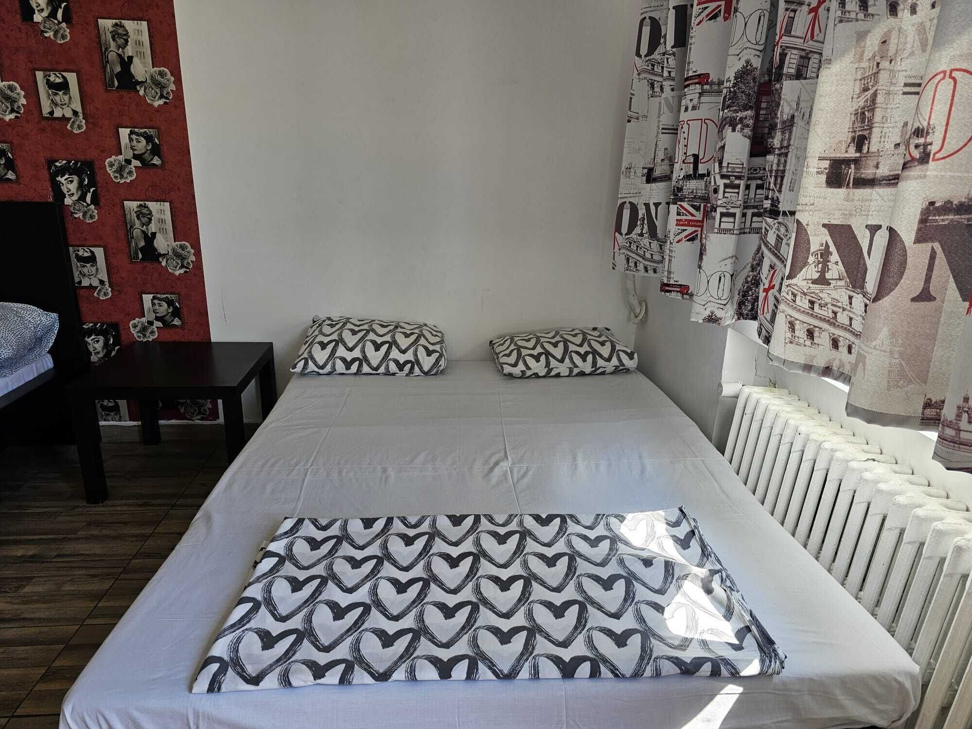 A bed with heart-patterned pillows, a red patterned wall, and a curtain with cityscape prints.