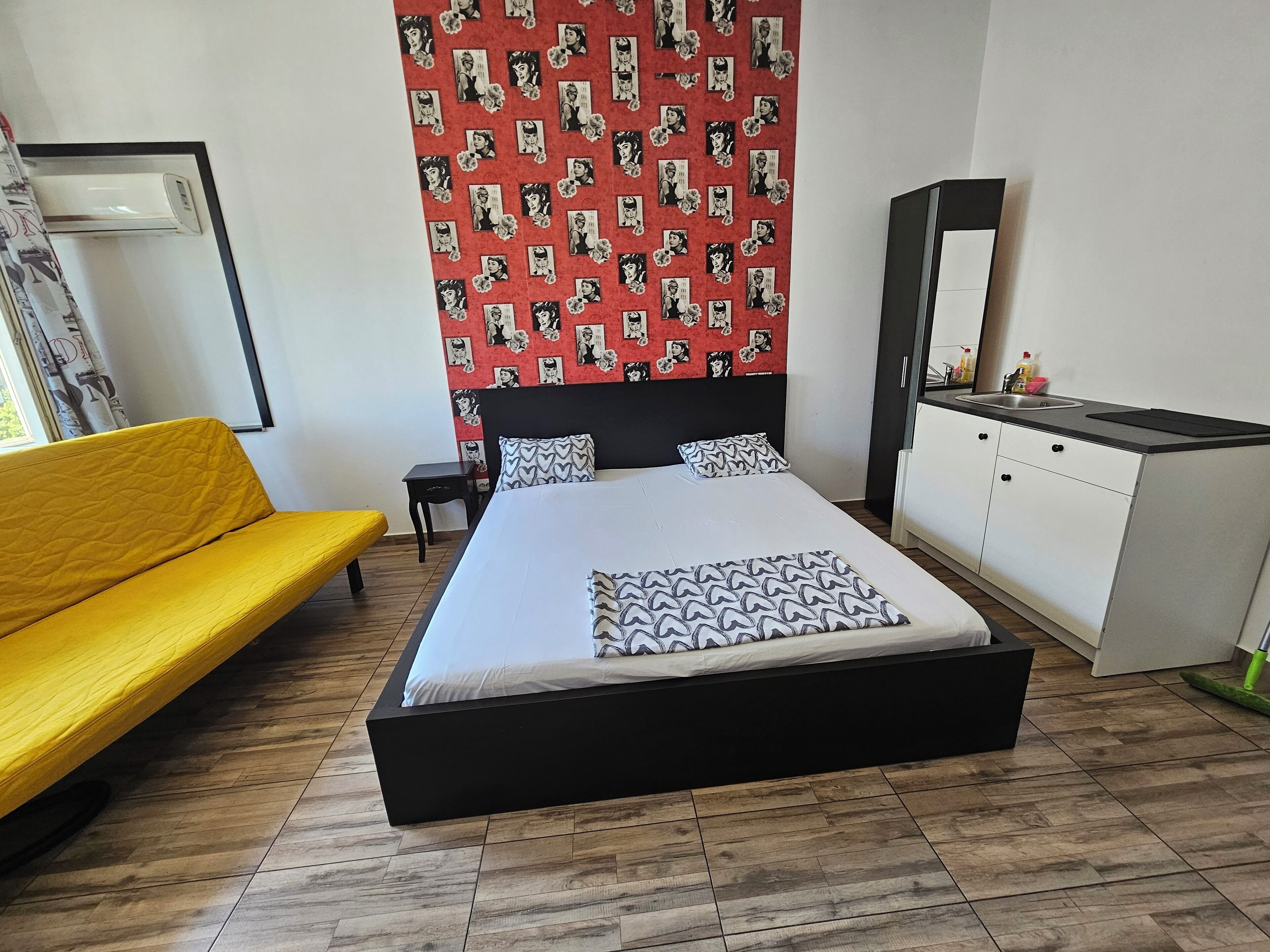A bedroom with a bed, a yellow sofa, a small bedside table, a dresser, and a mirror.