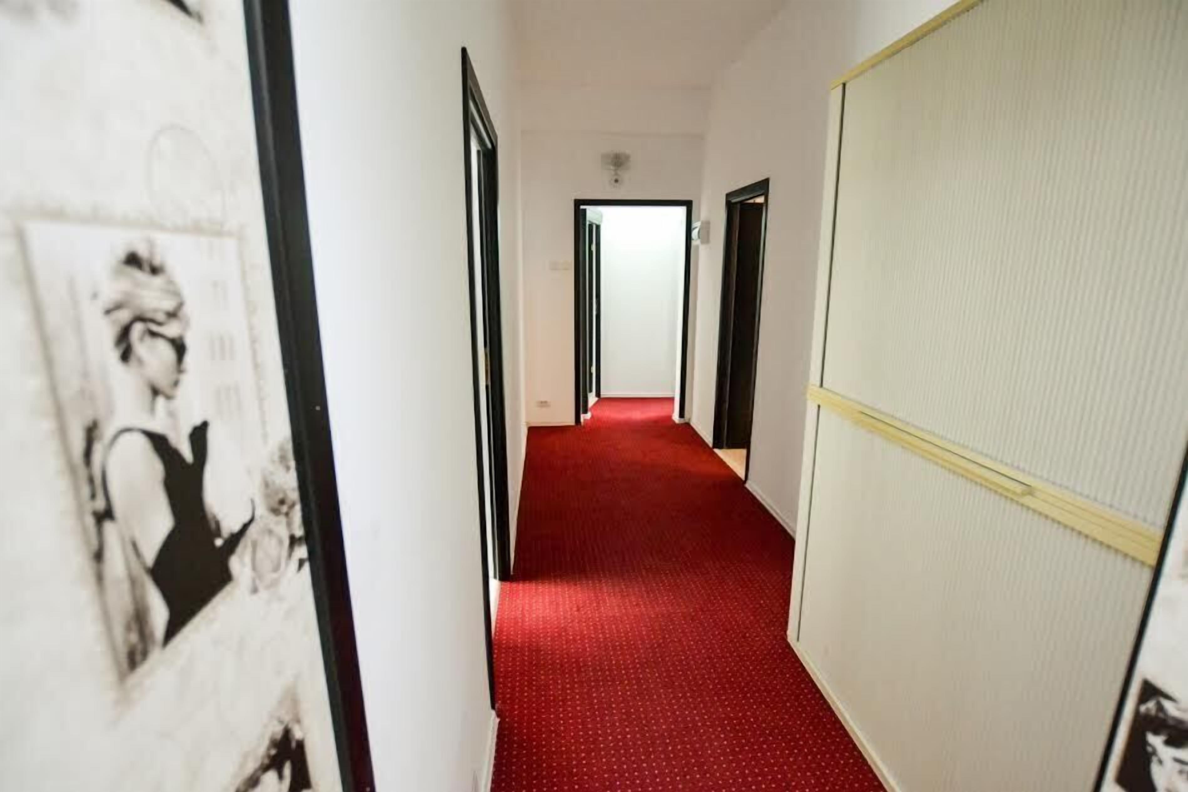 Un pasillo con alfombra roja, paredes blancas y puertas que dan a habitaciones.