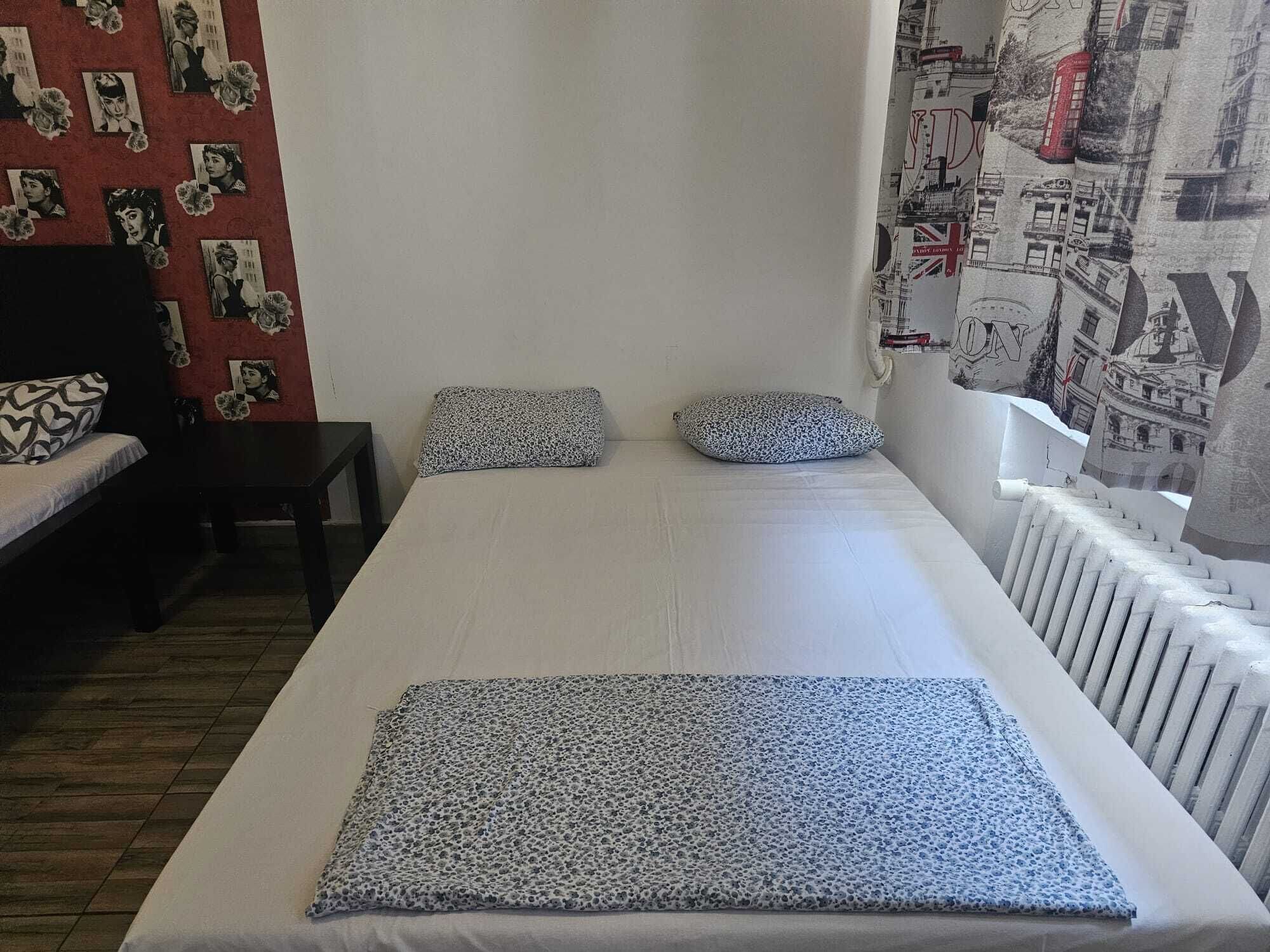 Una cama individual con un cojín estampado y una colcha, una mesita pequeña y una pared con carteles.