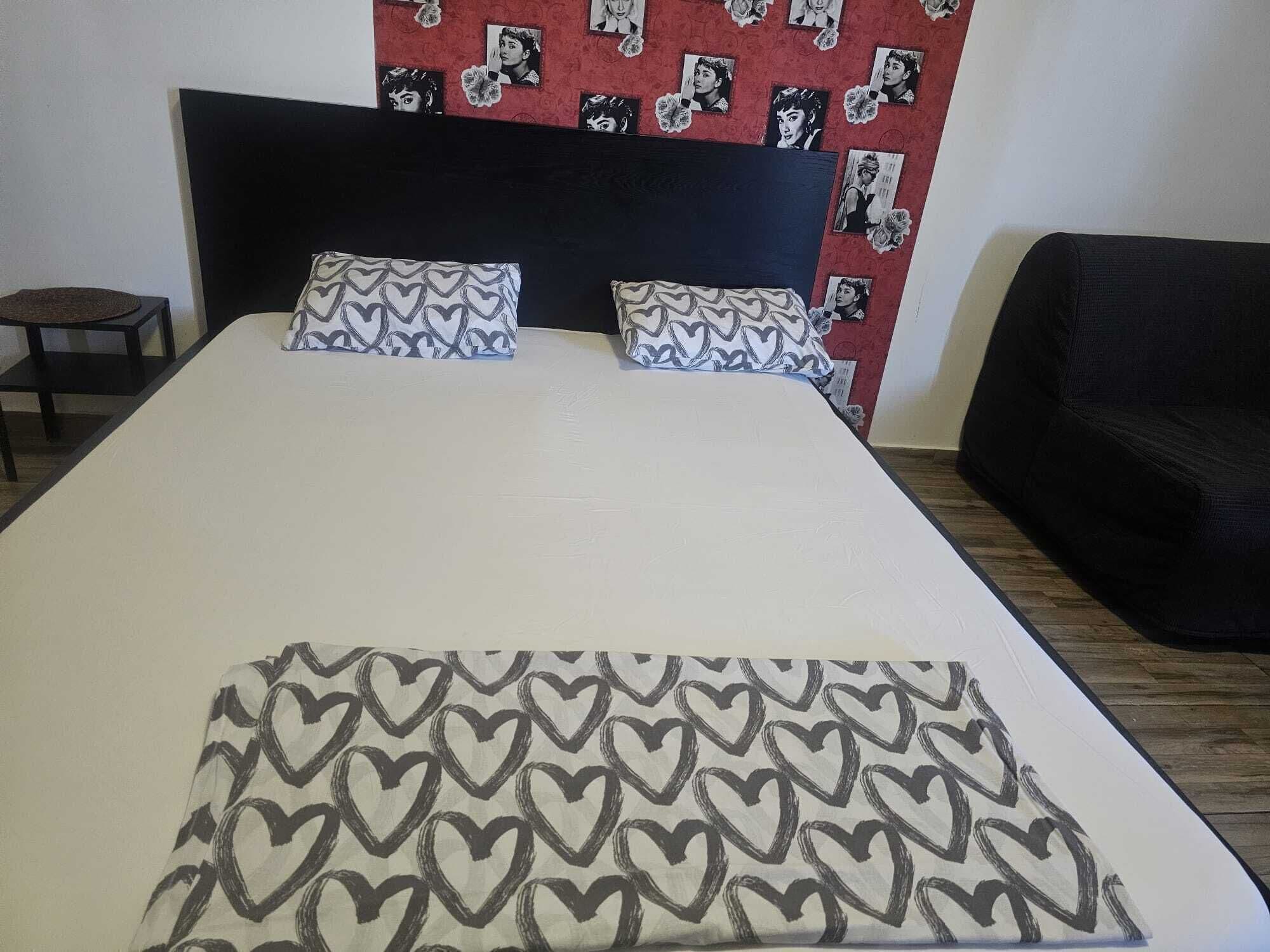 A bed with a patterned bedspread, two pillows with a heart design, and a wall with framed pictures.