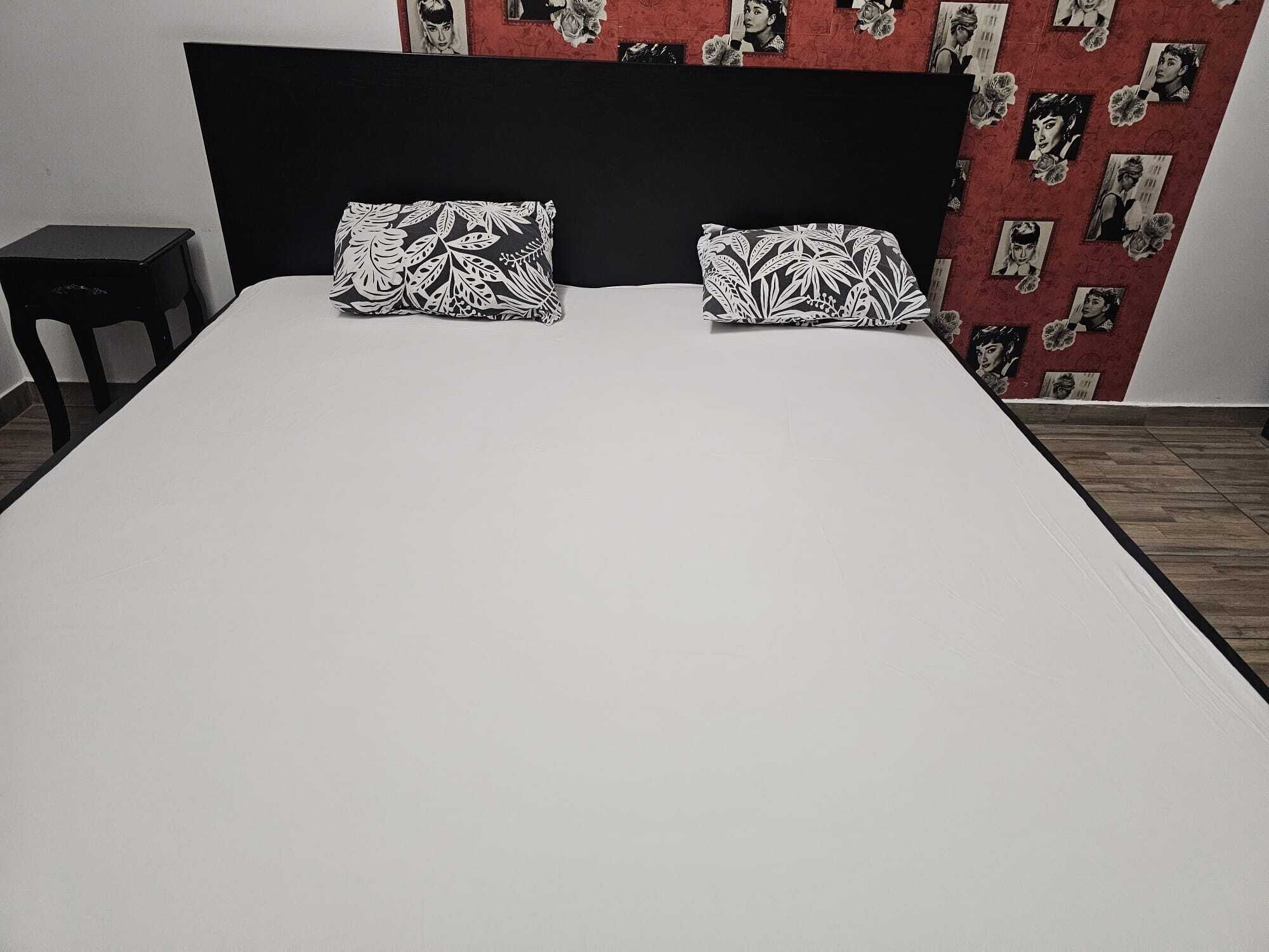 Una cama con cabecera negra, dos almohadas blancas con diseño de hojas y una pared roja con imágenes en blanco y negro.