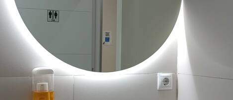Une salle de bain avec un lavabo blanc, un robinet noir, un miroir rond avec un éclairage intégré et un distributeur de gel hydroalcoolique.