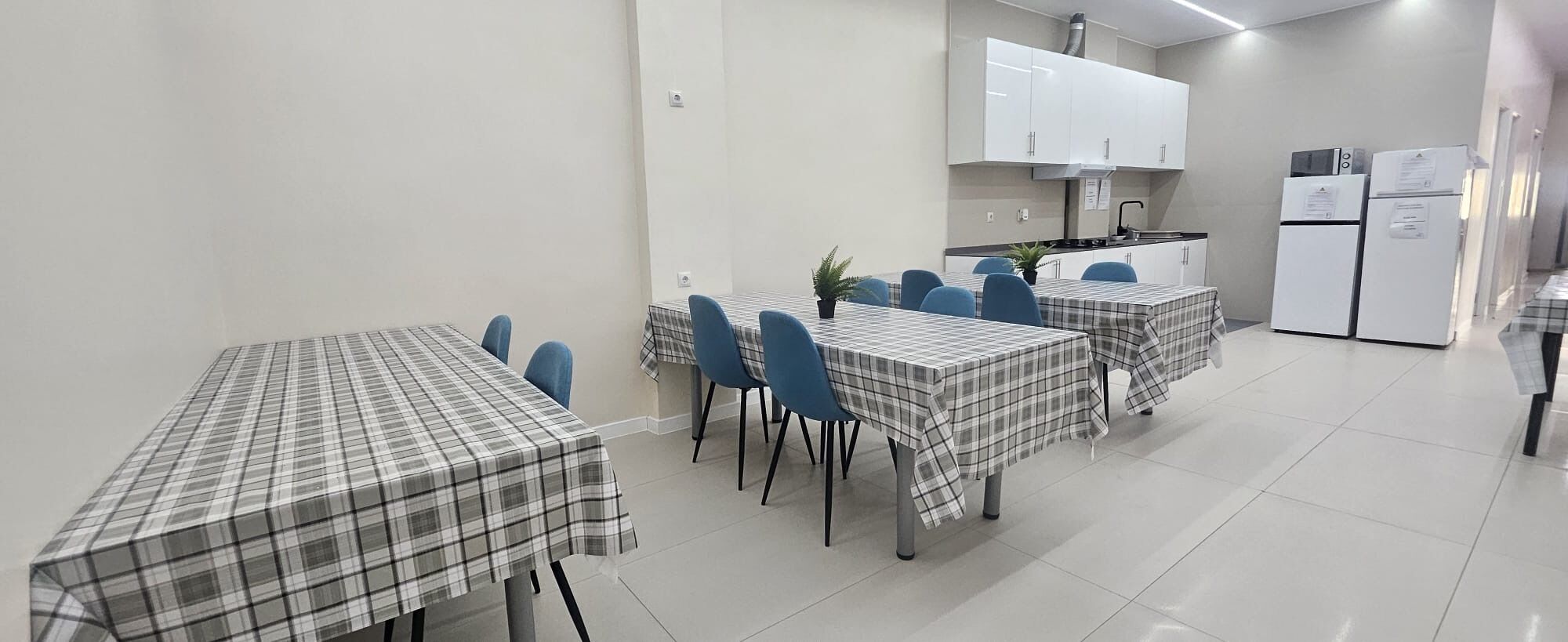 Cocina moderna con armarios blancos, microondas y refrigerador. Hay dos mesas de comedor con manteles escoceses y sillas azules.