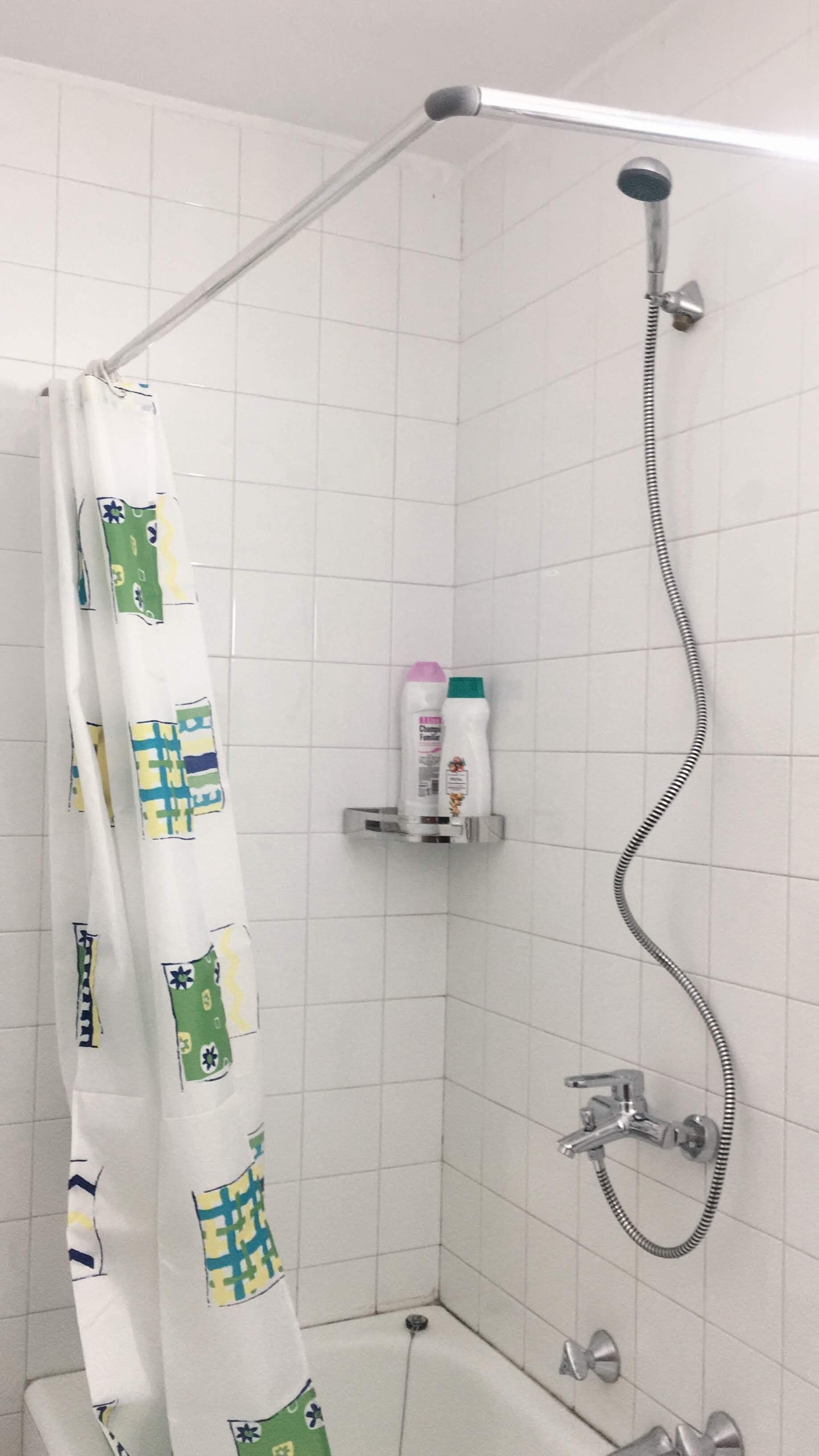 A shower area with a white tiled wall, a showerhead, a handheld shower attachment, and a shower curtain with a geometric pattern.