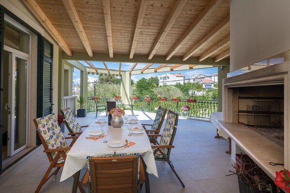 Eine überdachte Terrasse mit Esstisch, Stühlen und Grillbereich.