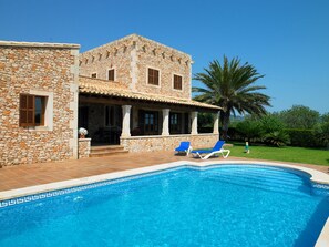 Una villa de piedra con piscina, dos sillones de descanso y una palmera.