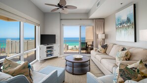 A spacious living room with a large TV, comfortable seating, a round coffee table, and a balcony with a view of the ocean. - Gulf Views ~ Balcony ~ Full Kitchen ~ Resort Pool (Miramar Beach)