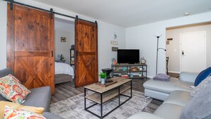 A living room with a sofa, a coffee table, and a TV. There is a sliding barn door and a white door in the background.
