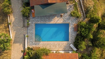 An aerial view of a rectangular swimming pool surrounded by stone tiles, with a tiled deck area and a covered patio.