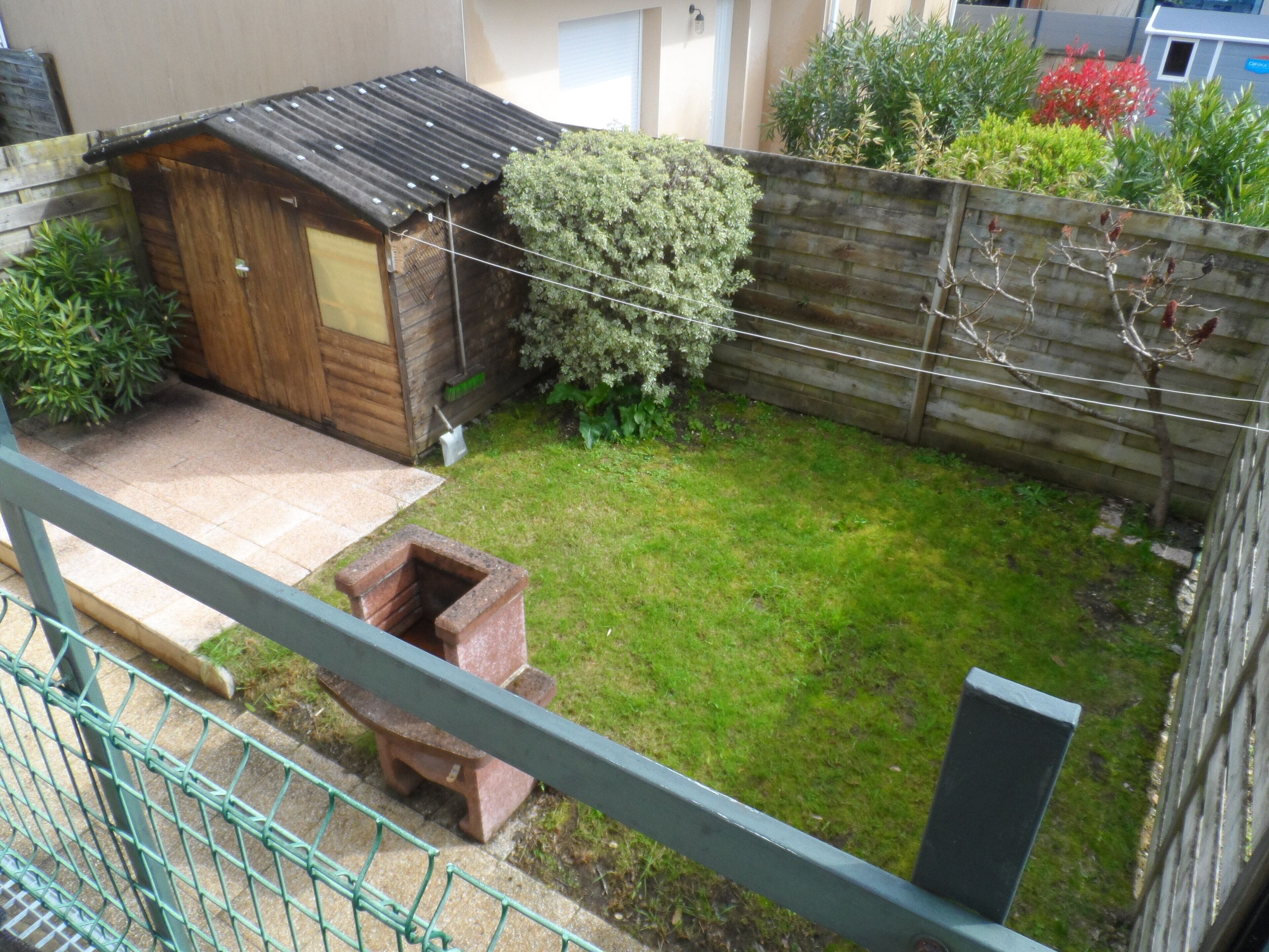 Een omheinde achtertuin met een houten tuinhuis, een bakkenplanter van baksteen en verschillende planten.