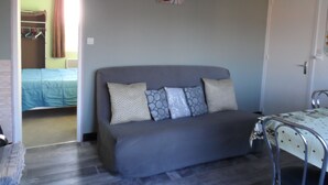Een kamer met een grijze bank, een metalen stoel, een bed met een blauwe sprei en een deur die naar een andere kamer leidt.