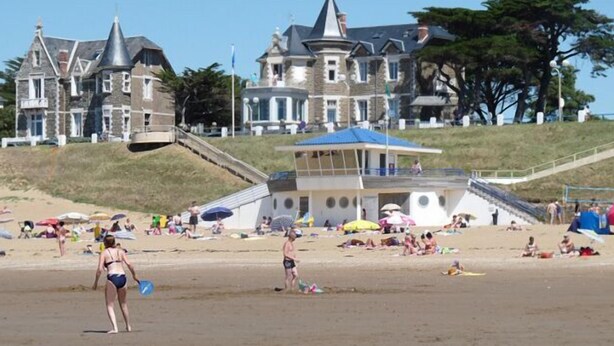 Een strand met mensen die zonnebaden en zwemmen, een redershut en een groot huis op de achtergrond.
