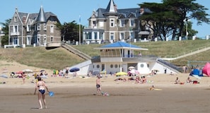 A beach with people sunbathing and swimming, a lifeguard tower, and a large house in the background.