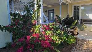 Ett hus med en täckt veranda, ett vitt spaljérstaket och en trädgård med rosa blommor.