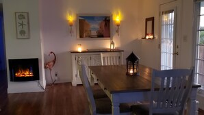 En matplats med ett träbord och stolar, en takfläkt, en öppen spis med en tänd brasa, väggmonterade lampor och en tavla ovanför bordet.