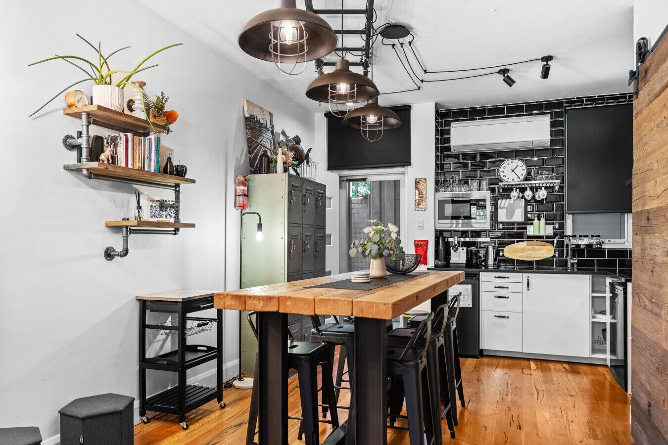 A modern kitchen with black cabinets, a wooden table, and bar stools. There is a hanging light fixture above the table.