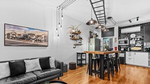 Et moderne køkken med en central ø, et spiseområde med et træbord og stole, en sort lædersofa og en køkkenbordplade med hårde hvidevarer.