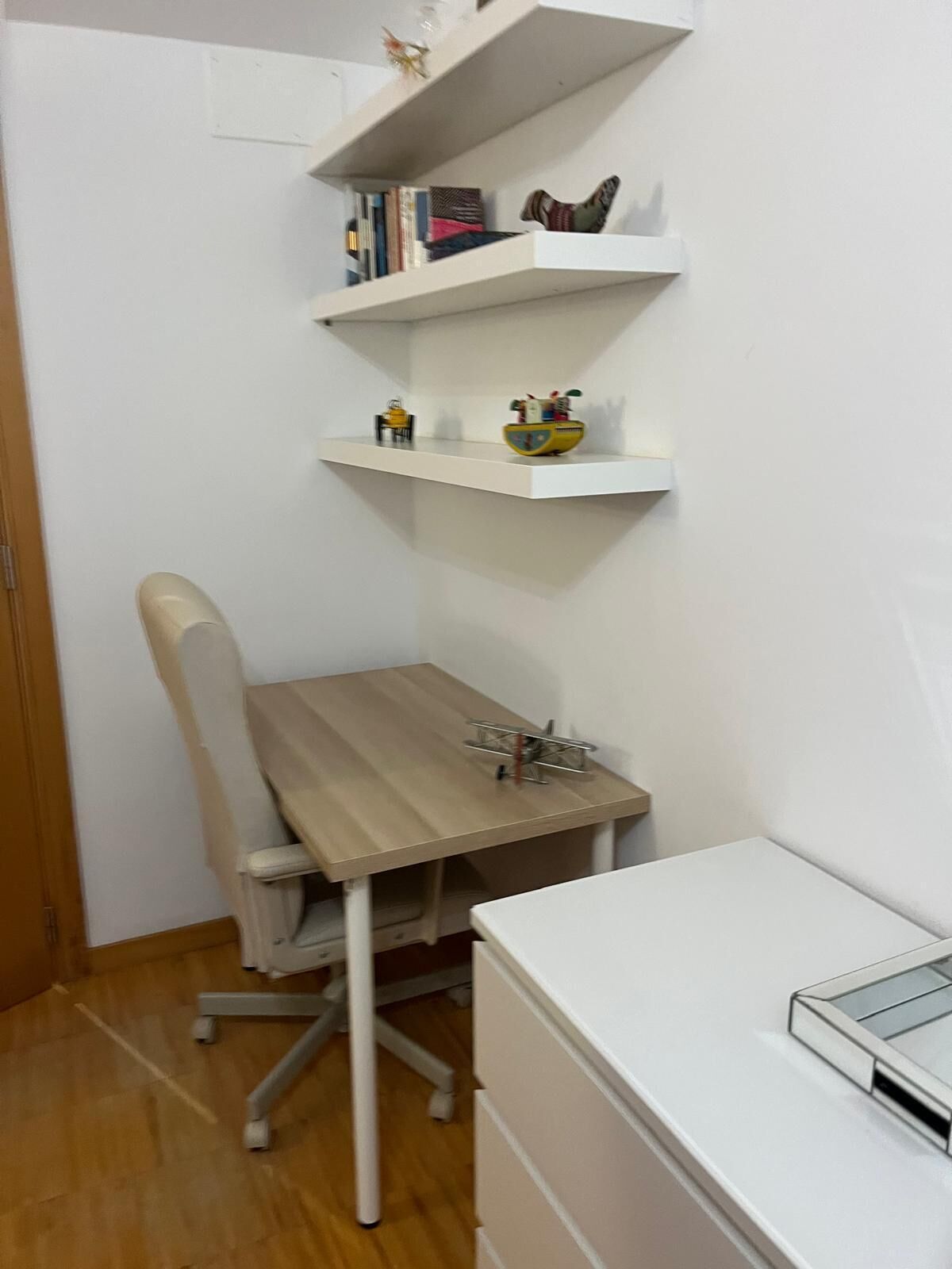 A corner room with a wooden desk, a white chair, a white dresser, and two white shelves with books and a toy car.