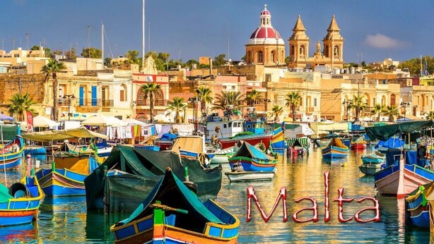 Ein Hafen mit zahlreichen bunten Booten an den Kais, eine markante Kirche mit Kuppel und das Wort "Malta" ist sichtbar.
