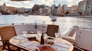 Ein Esstisch mit Geschirr, Gläsern und einer Flasche auf einem Bootsdeck mit Blick auf eine Stadt.
