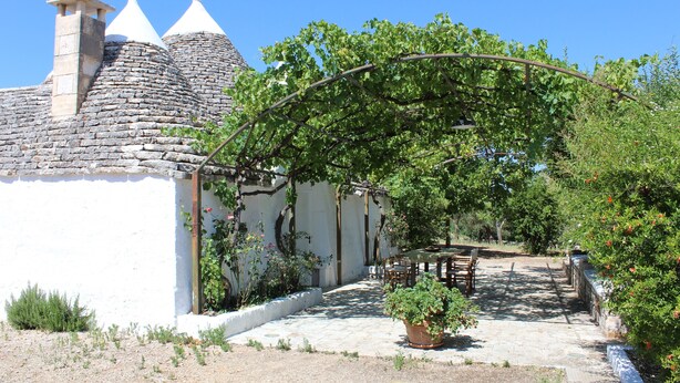 Een landelijk gebouw met een stenen dak, een witte muur en een tuin met een pergola.