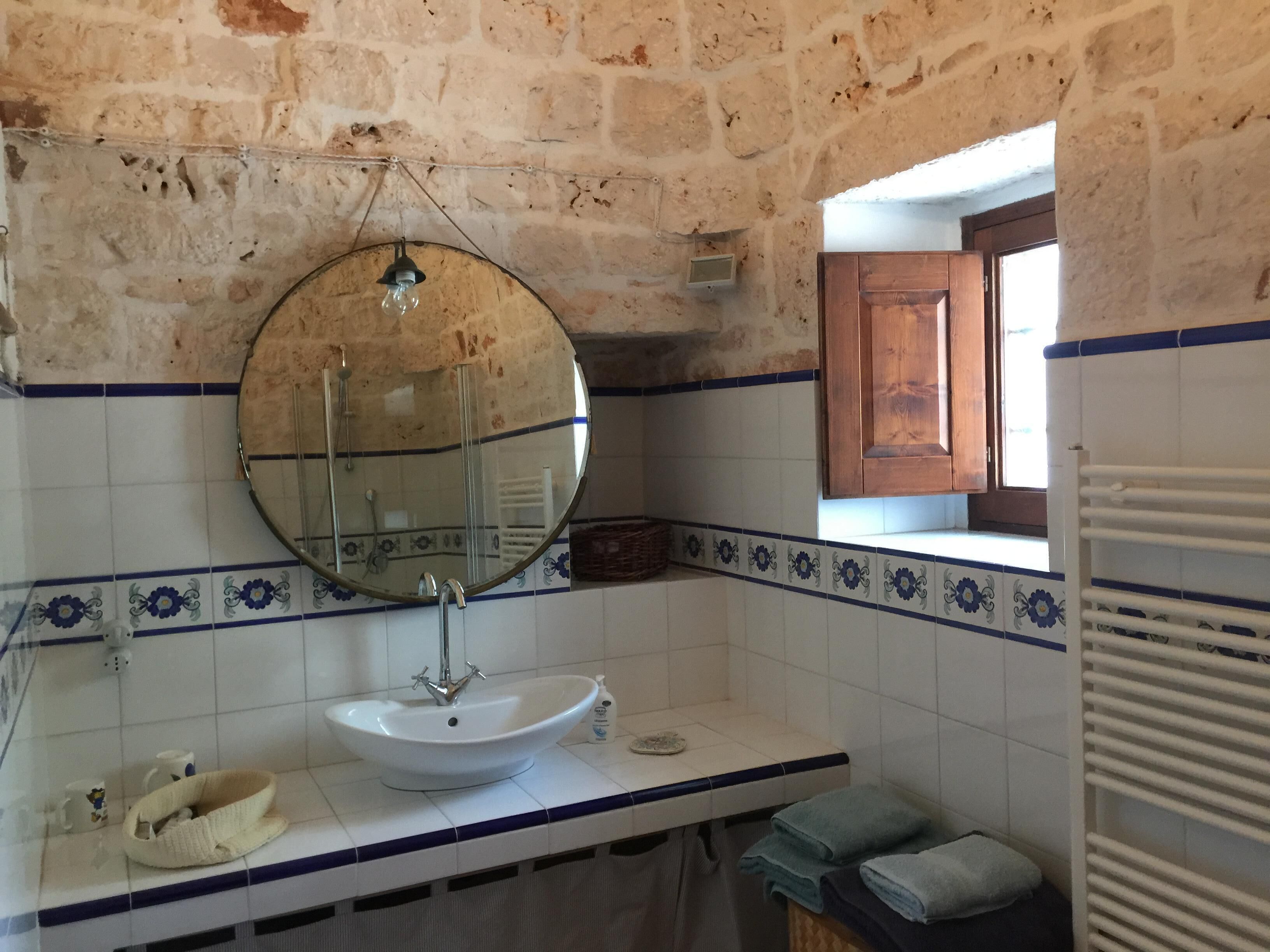 A bathroom with a white sink, a round mirror, and a window with a wooden door.