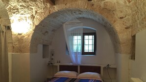 Een slaapkamer met een plafond van natuursteen, een bed met een beddekleed met dessin, een nachtkastje, een raam met uitzicht en een radiator.