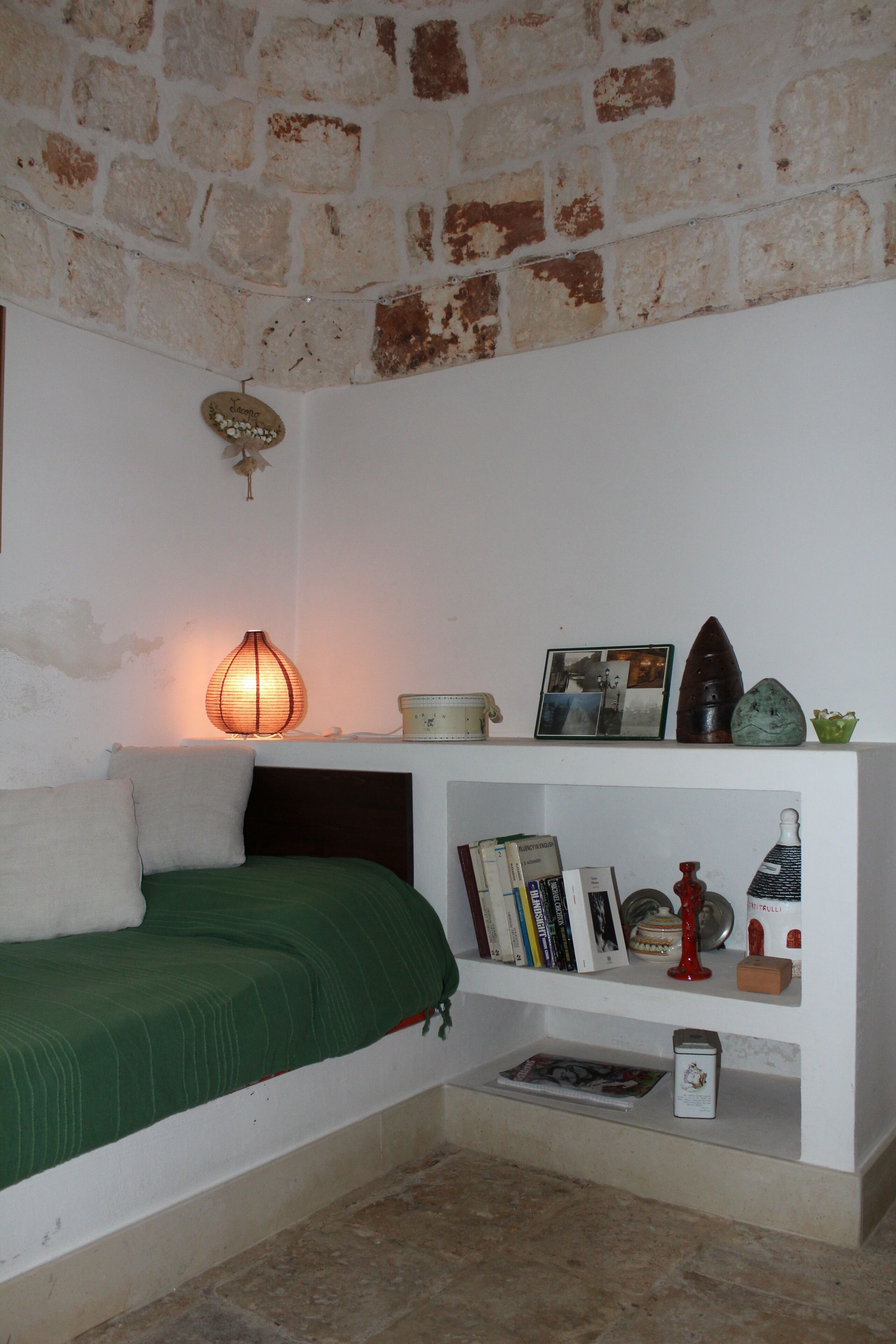 Una habitación con cama, una estantería con libros y adornos, una pared de piedra y una lámpara de pared.