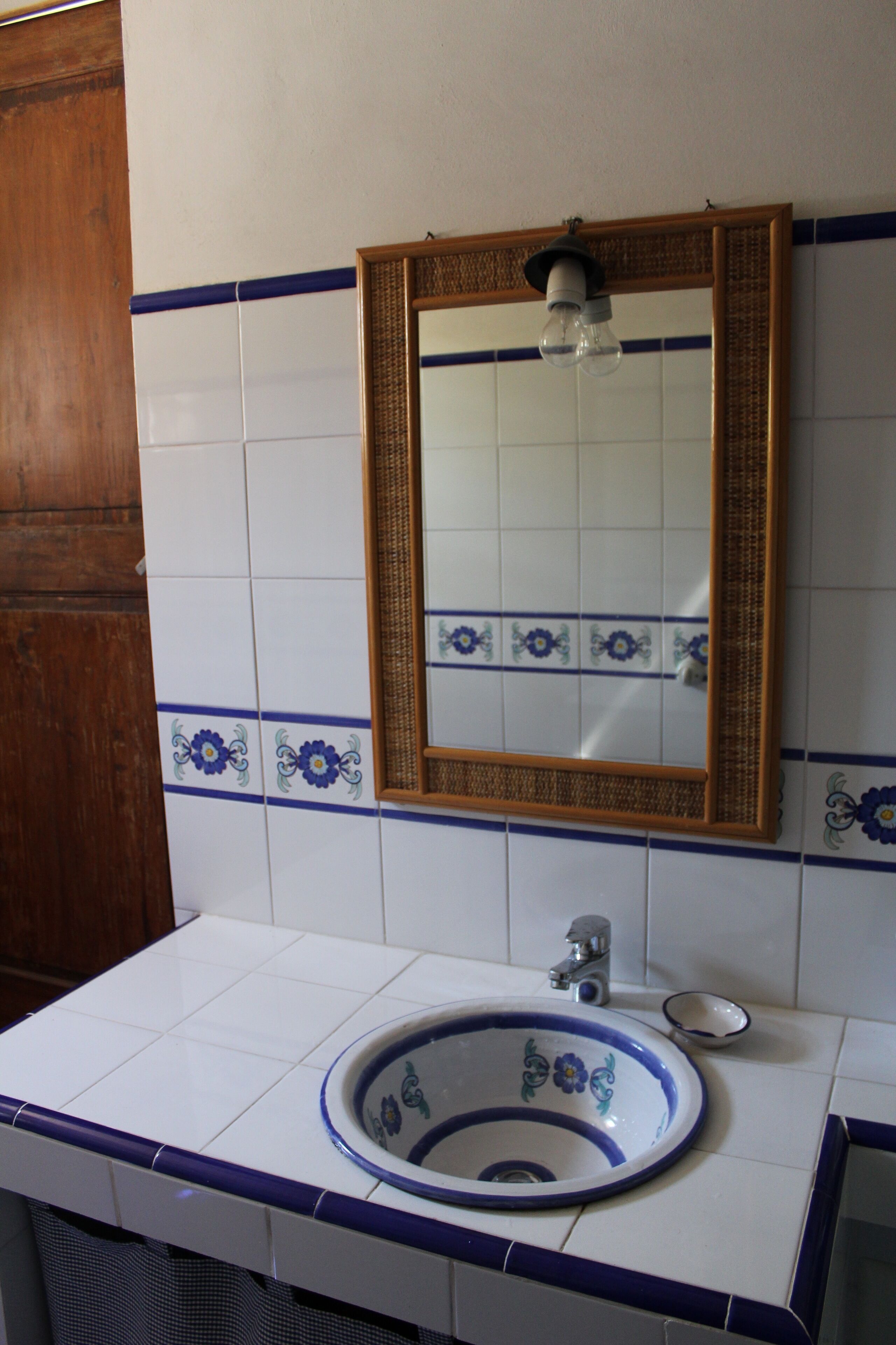 A bathroom with a white sink, blue and white tiles, a wooden door, and a mirror with a light fixture above it.