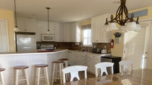 A kitchen with white cabinets, a central island with stools, a refrigerator, microwave, toaster, and a chandelier. - I LOVE BEACH MUSIC (Ocean Isle Beach)